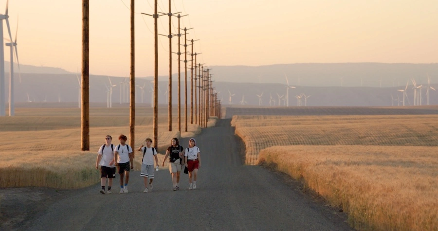 Immagine dal film Gasoline Rainbow: i cinque ragazzi camminano su una strada asfaltata tra campi di grano